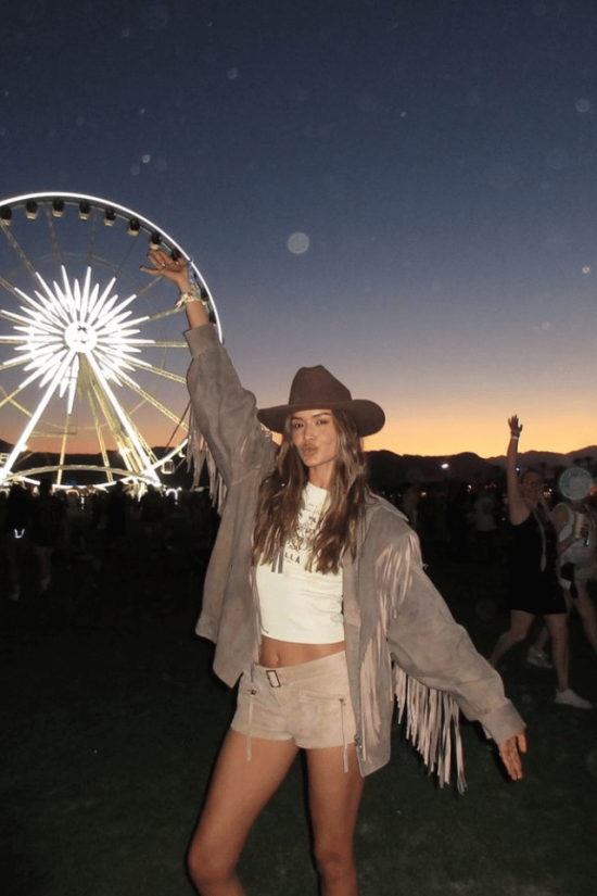 11 Country Concert Outfit Ideas woman in Country Concert Outfit Ideas with cowboy hat and fringe leather jacket in front of a ferris wheel at night.